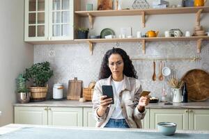 frustrado engañado mujer a hogar en cocina rechazado, incorrecto dinero transferir, Internet fraude, latín americano mujer triste participación banco crédito débito tarjeta y teléfono, utilizando en línea compras aplicación foto