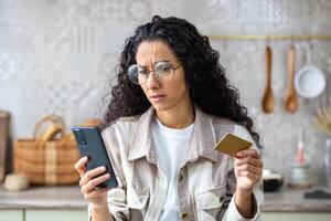 Frustrated cheated woman at home in kitchen rejected, wrong money transfer, internet fraud, latin american woman sad holding bank credit debit card and phone, using online shopping app. photo