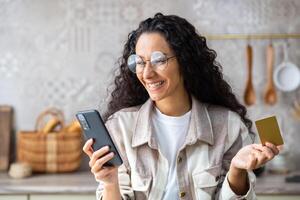 joven hermosa mujer en cocina con teléfono y banco crédito débito tarjeta en manos, sonriente Hispano mujer elige regalos y productos en en línea almacenar, en línea compras remotamente desde hogar. foto