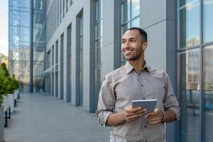 un empresario con un tableta computadora en su manos desde el fuera de de un oficina edificio, el hombre es sonriente y mirando lejos. un empleado en un camisa camina fuera de un oficina edificio, usos un aplicación foto