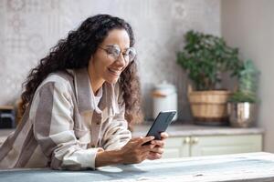 un joven hermosa mujer a hogar en cocina sonrisas contentamente, navega social redes, en línea Internet páginas, sostiene teléfono en r manos. Hispano con Rizado pelo , utilizando un aplicación en teléfono inteligente foto