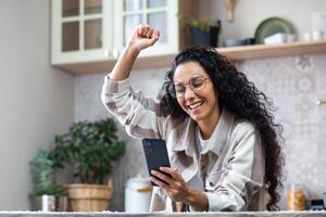Joyful woman at home celebrating victory and successful achievement results, winner looking at phone received offer online, Hispanic woman inside home in kitchen. photo