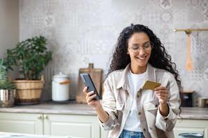 joven hermosa mujer en cocina con teléfono y banco crédito débito tarjeta en manos, sonriente Hispano mujer elige regalos y productos en en línea almacenar, en línea compras remotamente desde hogar. foto