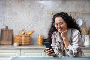 un joven hermosa mujer a hogar en cocina sonrisas contentamente, navega social redes, en línea Internet páginas, sostiene teléfono en r manos. Hispano con Rizado pelo , utilizando un aplicación en teléfono inteligente foto