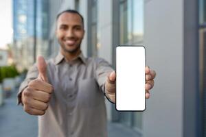 Young successful man showing white phone screen to camera, businessman recommending app, looking happily at camera, outside office building. photo
