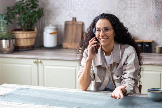 joven hermosa mujer a hogar en el cocina hablando en el teléfono felizmente, exitoso latín americano mujer en el cocina cerca arriba con Rizado pelo y lentes. foto