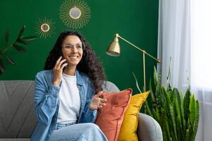 un alegre mujer se involucra en un conversacion en su teléfono inteligente mientras sentado cómodamente en su vibrante vivo habitación, rodeado por decorativo plantas de interior foto