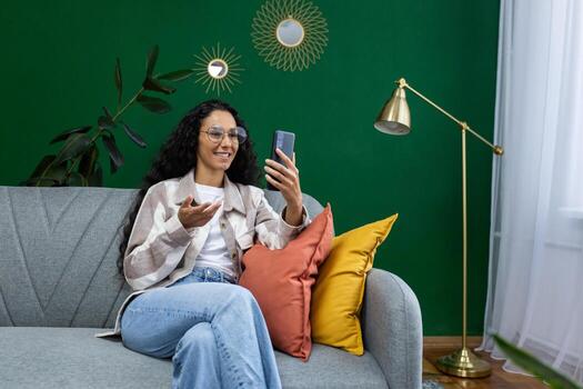 joven hermosa mujer hablando en llamada utilizando aplicación en teléfono inteligente, Hispano mujer sentado en sofá en vivo habitación, sonriente a interlocutor. foto