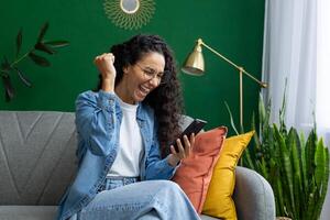 un extático mujer en anteojos, vistiendo un mezclilla chaqueta, es visto levantamiento su puño en victoria mientras mirando a su teléfono inteligente, sentado cómodamente en un sofá adornado con vistoso almohadas foto