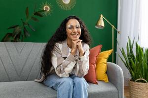un alegre mujer en casual ropa disfruta un cómodo momento sentado en un gris sofá, rodeado por vibrante plantas de interior y elegante hogar decoración. foto