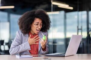 African American businesswoman has sore throat while working in office, uses throat spray, has cold, cough. photo