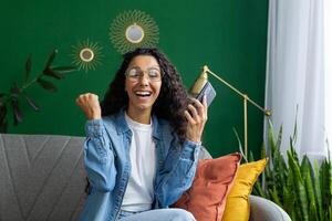 esta imagen capturas un alegre mujer celebrando un personal victoria o recepción bueno Noticias en su teléfono inteligente, mientras sentado cómodamente en un elegantemente decorado habitación. foto