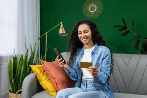 Young beautiful joyful woman sitting on sofa at home with phone and bank credit debit card in hands, Hispanic woman in living room at home smiling choosing gifts, shopping online in online store. photo