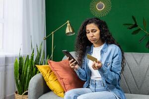 Sad cheated woman holding bank credit debit card and phone, hispanic woman rejected fund transfer, insufficient account balance, sitting on sofa in living room at home. photo