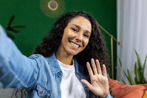 cámara vista, mujer a hogar en sofá mirando dentro teléfono cámara sonriente y hablando con amigos utilizando teléfono inteligente aplicación, ondulación mano gesto de saludo, y tomando selfie foto, en vivo habitación. foto