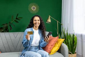 Portrait of joyful woman at home on sofa in living room holding bank credit card and phone, Hispanic woman looking at camera, using online shopping app. photo