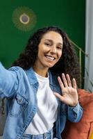 vertical disparo, mujer a hogar en sofá mirando a teléfono cámara sonriente y hablando con amigos utilizando teléfono inteligente aplicación, ondulación saludo gesto y tomando selfie foto, en vivo habitación. foto