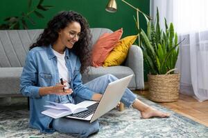 Young beautiful woman sitting on the floor at home in the living room, student studying remotely, using laptop to watch webinar and course, online call consultation with teacher. photo