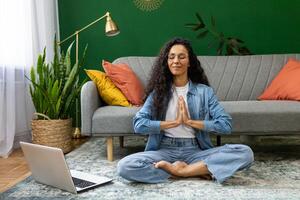 Young woman resting meditating in lotus pose sitting on floor at home in living room cinema, Hispanic woman with laptop doing online group yoga in training class remotely. photo