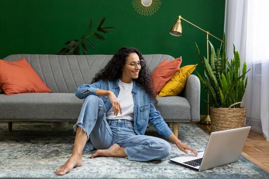 alegre exitoso mujer trabajando en ordenador portátil de forma remota, Hispano mujer sentado en alfombra en piso en vivo habitación, felizmente mecanografía en teclado. foto