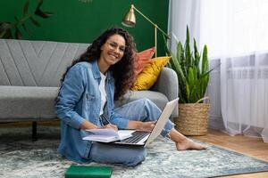 Portrait of young female student studying at home, Hispanic woman smiling and looking at camera, sitting on floor in living room with laptop, online learning, writing data in notebook. photo