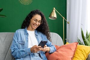 alegre joven mujer en lentes y mezclilla disfrutando su teléfono inteligente en un acogedor sofá con vistoso almohadas foto
