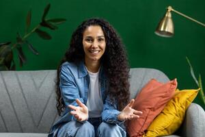 Young beautiful woman at home on sofa looking into web camera, talking with friends and colleagues remotely, smiling with satisfaction and gesturing with hands using call on computer. photo