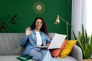 alegre mujer con lentes ondulación mientras participación un computadora portátil, sentado en un sofá con vistoso cojines y interior plantas. foto