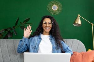 Cheerful woman in glasses and denim waving hello during a call at home, with a green wall and stylish decor. photo