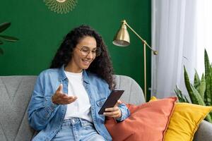 contento mujer en lentes utilizando teléfono inteligente en sofá con vistoso almohadas, sensación alegre en un acogedor hogar ajuste. foto
