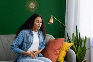 Surprised woman sitting on couch with hand on stomach, expressing shock or discomfort at home with green decor. photo