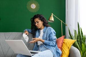 en línea consulta con un psicólogo, un psicoterapeuta, un llorando mujer sentado en un sofá en un vivo habitación con un computadora portátil, hablando remotamente con un doctor, utilizando un llamar. foto