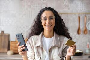 Happy hispanic woman holding phone and card, online shopping, internet payment, smiling and looking at camera. photo