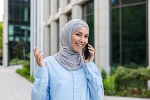 Businesswoman in hijab talking on the phone standing in front of the office, important negotiations, looking at the camera. photo