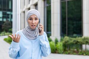 Sad Muslim female worker in hijab talking on the phone, standing on the street, spreading her hands, looking at the camera. photo