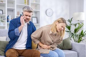 Worried husband calls an ambulance for his wife who has a stomach ache, husband calls on the phone while sitting on the sofa. photo