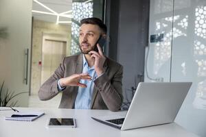 A professional businessman in a modern office setting communicates over the phone while gesturing actively, with a laptop and tablet in front of him. photo