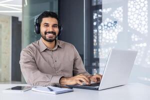 Carefree middle eastern guy in wireless headset typing on computer keyboard in modern office interior. Successful worker practising multitasking while listening to audiobook and answering emails. photo