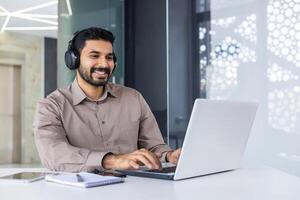 Young successful Indian programmer at workplace inside office, man with headphones smiling satisfied, coding new software using laptop at table, businessman in business shirt successful, satisfied photo