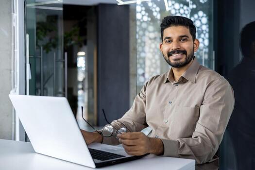 retrato de contador financiero a lugar de trabajo, empresario con ordenador portátil sonriente y mirando a cámara dentro oficina a lugar de trabajo, hombre en camisa contento con logro resultados y exitoso informes. foto