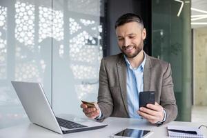 A smiling businessman in a modern office uses a phone and credit card for online transactions while working on a laptop. photo