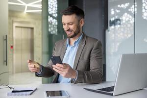 sonriente empresario en moderno oficina haciendo en línea pago utilizando crédito tarjeta y teléfono inteligente, retratar facilitar de digital actas. foto