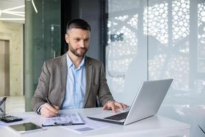 A focused business man reviewing documents while working on his laptop in a modern office setting, with natural light backlighting the scene. photo