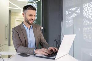 confidente y alegre empresario utilizando un ordenador portátil para trabajo en un brillante contemporáneo oficina ajuste. foto