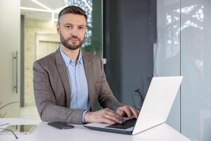 A focused professional man in a suit working intently on a laptop in a modern office setting, exuding a serious demeanor. photo