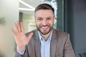 Friendly professional man in a suit waving and smiling at the camera, appearing to be on a conference call in an office setting. photo