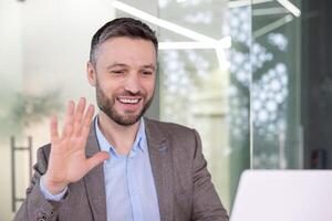 Cheerful professional man in a smart suit waving his hand while having a conference call in a modern office setting. photo