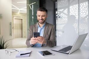 alegre sonriente satisfecho empresario mecanografía texto mensaje en teléfono, hombre en negocio traje hojeada social medios de comunicación y utilizando aplicación en teléfono inteligente, sentado dentro oficina con ordenador portátil a lugar de trabajo. foto