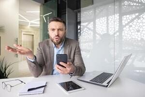 Confused businessman with phone at desk expressing frustration, surrounded by tech gadgets in a modern office setting. photo
