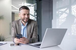 A young male businessman sits at a desk in the office, holds a phone in his hands and communicates via call on a laptop. photo